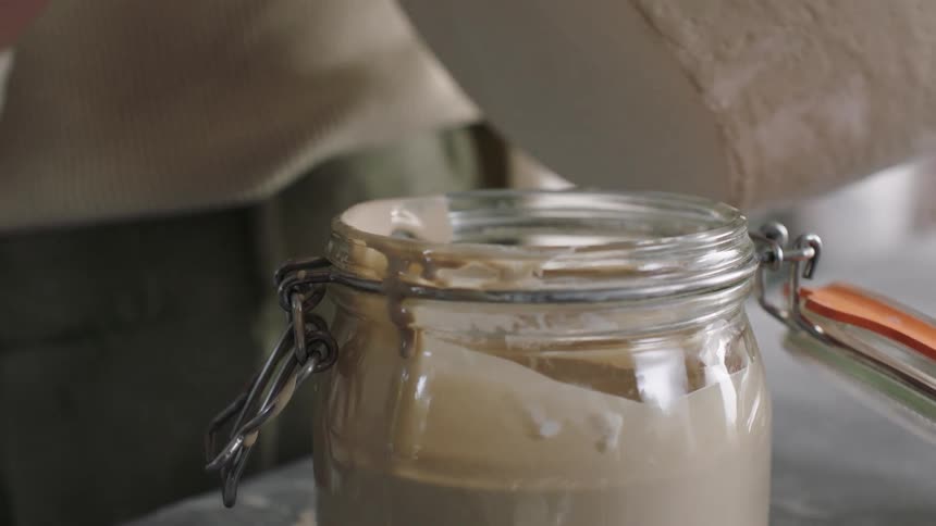 A person uses a wooden brush to stir the ceramic glaze in a glass jar before applying the liquid suspension of powdered minerals onto a pottery piece