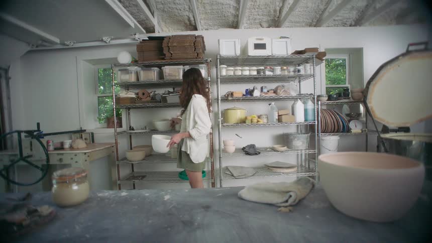 Young adult woman in a pottery studio carrying ceramic bowls and placing them on a shelf next to similar items indoors during day time