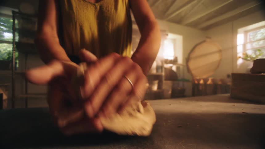 A ceramist in a yellow tank top firmly kneads a lump of clay on a dusty table preparing the material for her pottery project inside a sunlit studio