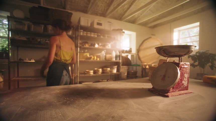A woman in a sunlit pottery studio briefly weighs and prepares a lump of clay on a dusty table before walking toward shelves in the background to continue her work