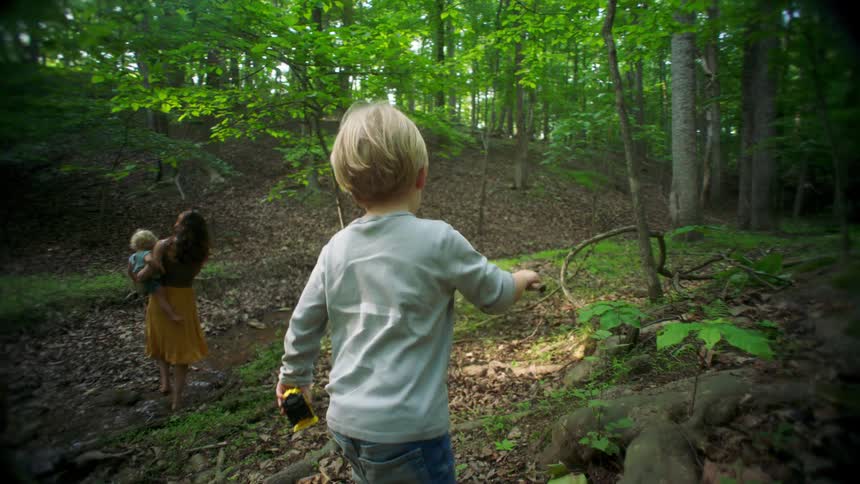Child walks through a forested area while a woman carries another child in the background