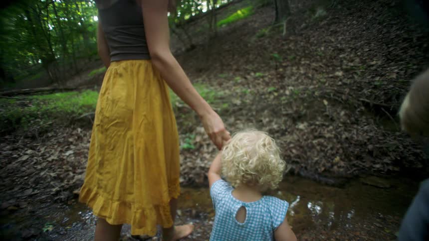 Woman holds a childs hand while another walks ahead near a stream in a forested area