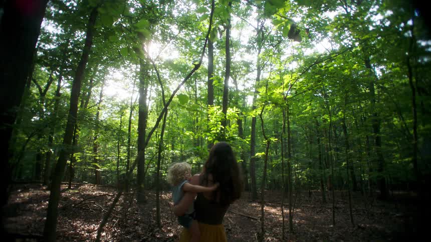 An adult woman carrying a blonde child in her arms is walking with her back to the camera in a green forest