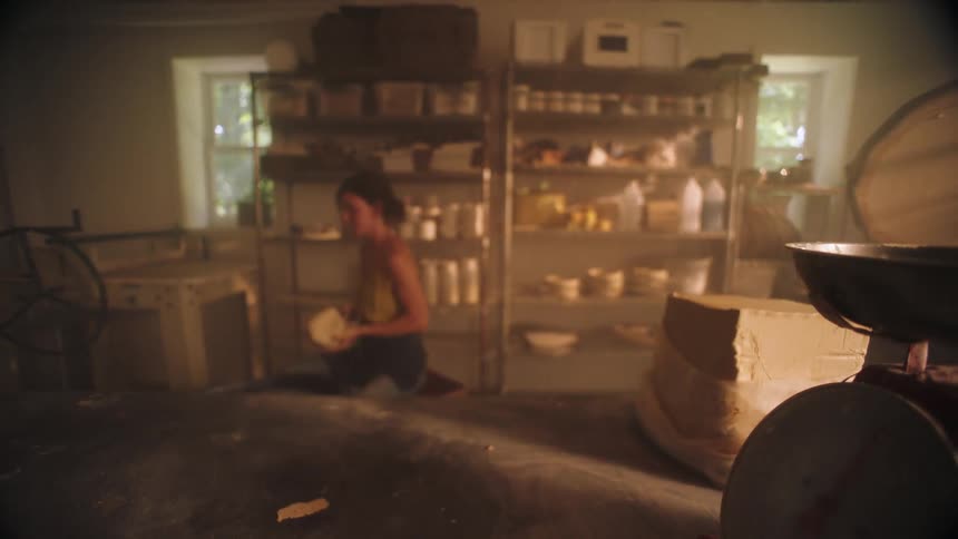 An artist kneels on the floor of her warm hazy studio shaping a piece of clay as sunlight drifts through the windows