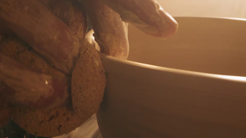 A potters hands covered in clay skillfully shape the rim of a large clay bowl spinning on a pottery wheel under warm lighting