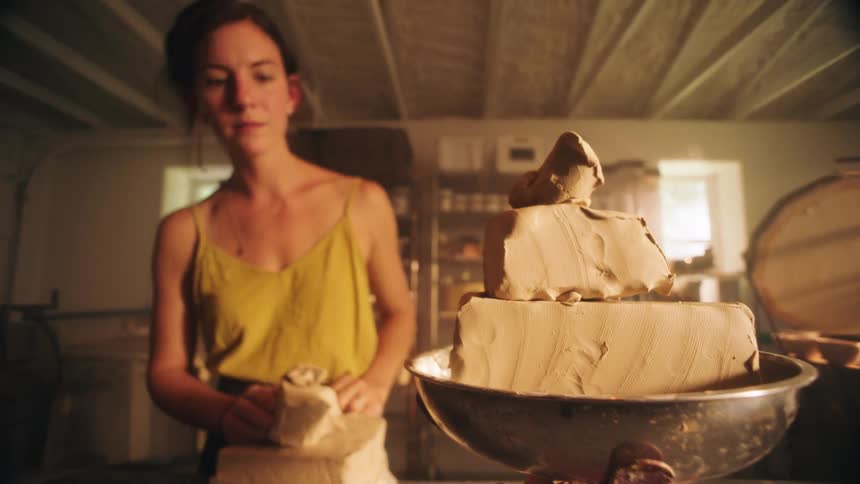 In a sundrenched pottery studio a focused artist stands before a vintage metal scale carefully tearing and adding chunks of raw clay to the weighing pan to measure a precise amount