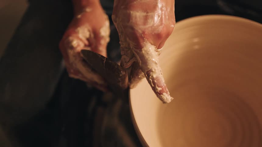A potters clay covered fingers and a tool smooth the rim of a large clay bowl spinning on the wheel