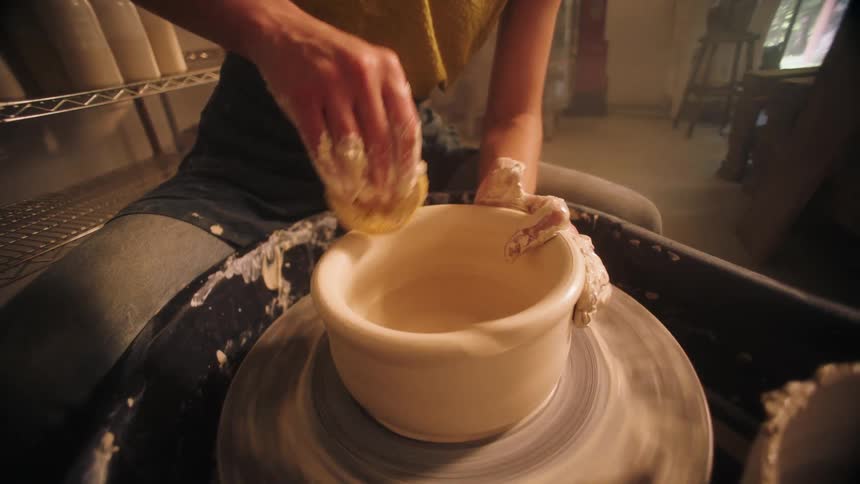 From a close perspective an artist uses a sponge to shape the inside of a wet clay bowl as it spins rapidly on a pottery wheel