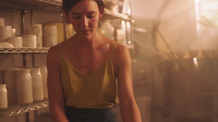 A young woman potter in a yellow tank top and apron prepares a lump of clay on a wheel in a dusty dimly lit studio filled with ceramic ware on metal shelves