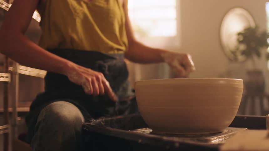 A woman potter wearing a yellow top and dark apron works on a large clay bowl centered on a spinning pottery wheel in a sunlit studio