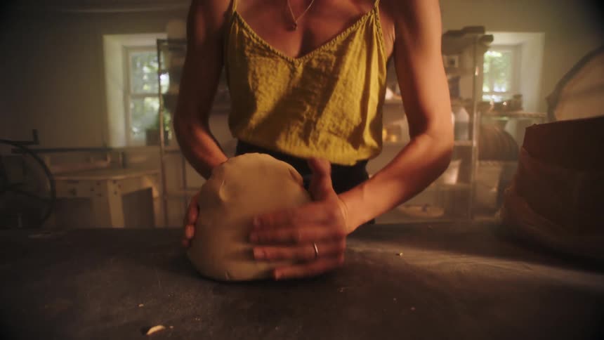 Bathed in warm hazy sunlight a focused artisan in a yellow tank top rhythmically kneads a large mound of clay on her workbench to prepare it for sculpting