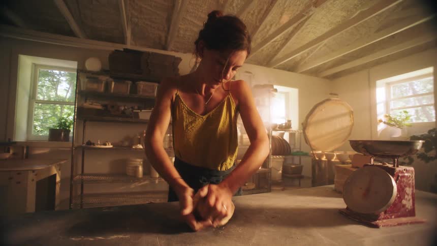 A woman stands centered at a dusty table in a brightly lit pottery studio intently kneading a lump of clay with both hands preparing the material