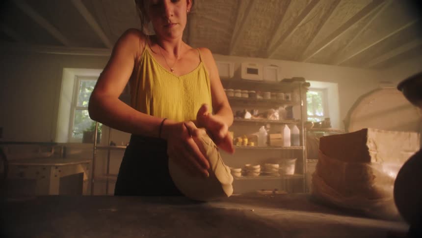 A woman potter wearing a yellow tank top and apron kneads a large block of clay on a dusty table in a dimly lit sunlit pottery studio
