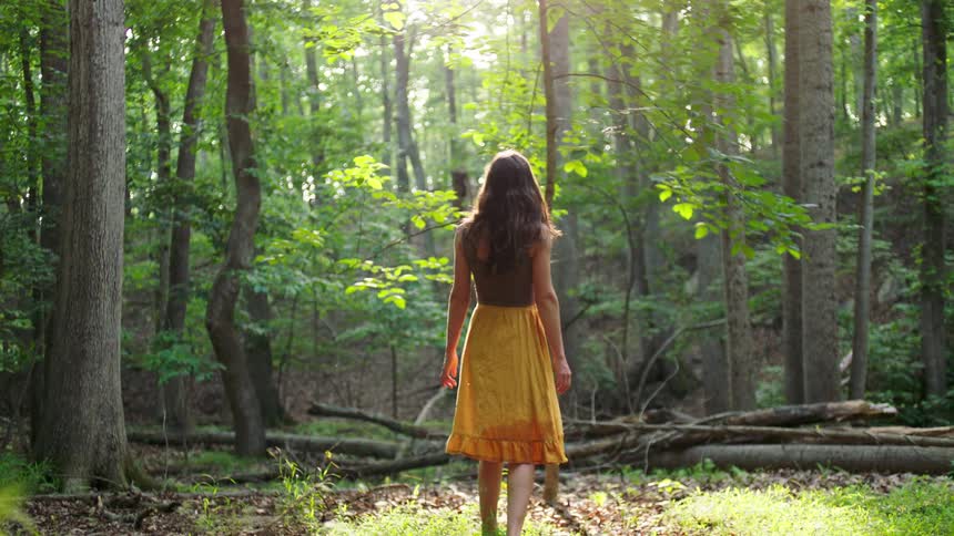 A woman with long hair wearing a brown top and yellow skirt walks through a lush green forest with sunlight filtering through the dense canopy