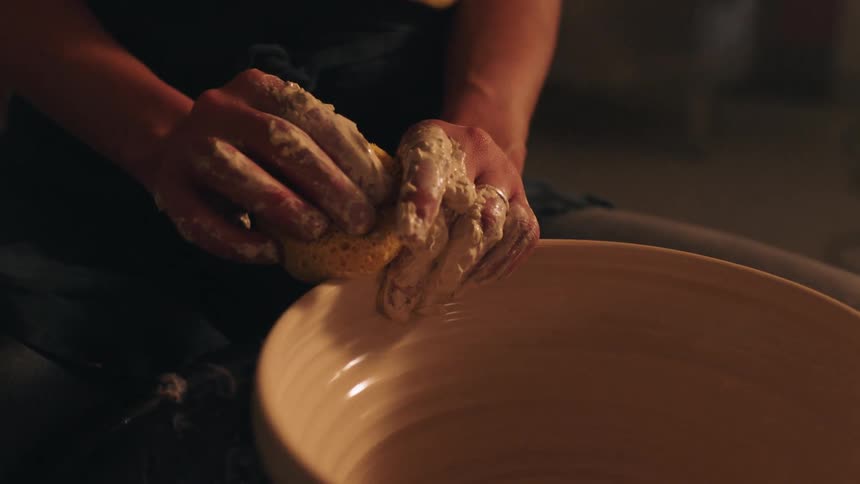 A potter reaches into a basin to soak a sponge then brings the dripping tool to the spinning clay bowl to moisten and shape its walls