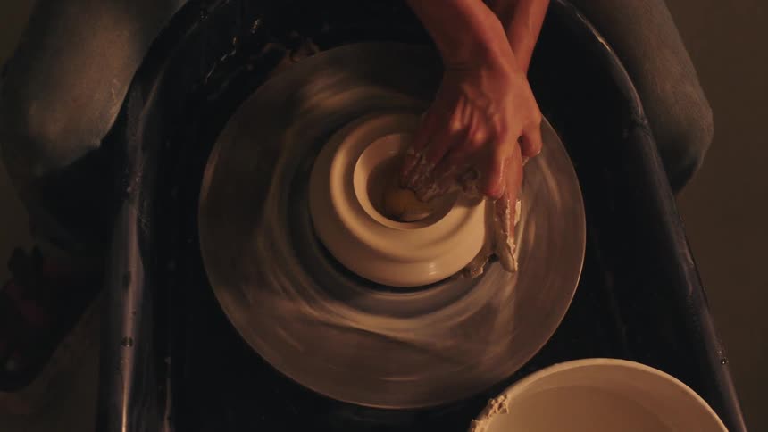 A close view from above shows a woman potter using a yellow sponge to shape a piece of white clay spinning on a pottery wheel in a dark studio