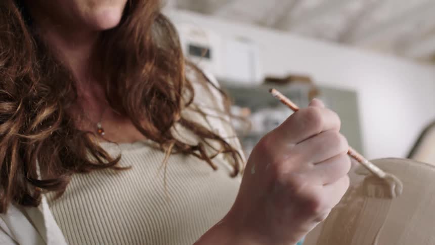 Hand applies coating to an unfired ceramic piece using a fine brush