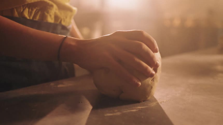 A woman in an apron and yellow top is captured in a warm hazy light repeatedly picking up and slapping a lump of clay to prepare it for pottery eventually placing the clay down on a table surface