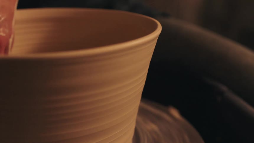 A potters hand gently presses a wet sponge against the rim of a spinning clay vessel smoothing the edge as it rotates rhythmically in the warm dim light