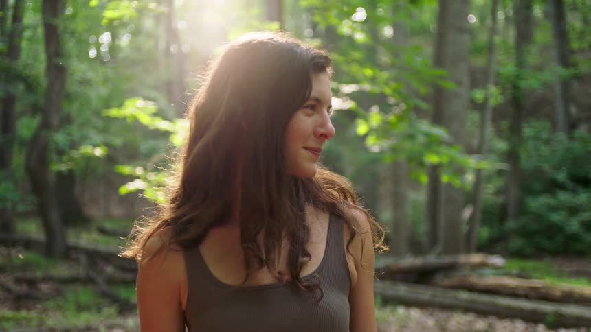A woman with long brown hair stands in a forest looking to her right while bright sunlight backlights her with the natural woodland environment surrounding her