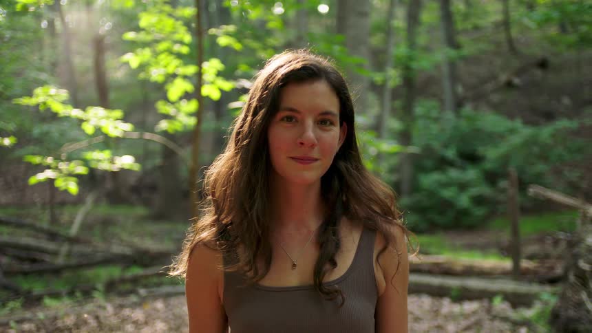 Woman with wavy brown hair in sunlit forest wearing sleeveless top and necklace