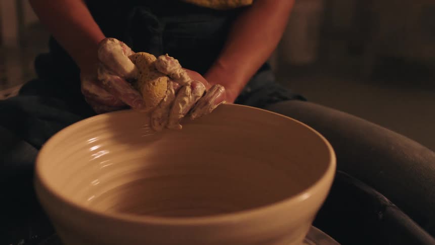 A person sitting at a pottery wheel works with their hands and a small yellow tool to sculpt the interior of a large spinning clay bowl resulting in defined horizontal texture rings