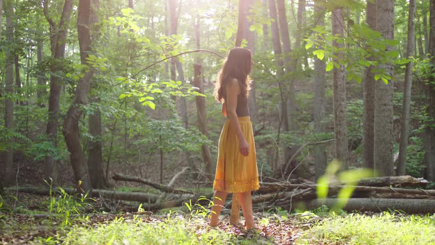 Sunlight streams through the tall trees as a woman in a flowing yellow skirt walks gently through the forest pausing to look around in the warm hazy glow