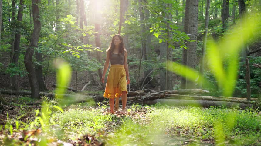 A woman barefoot in a yellow skirt and dark top stands alone in a sundrenched forest clearing surrounded by tall trees and lush green grass slowly turning to look up at the bright sunlight
