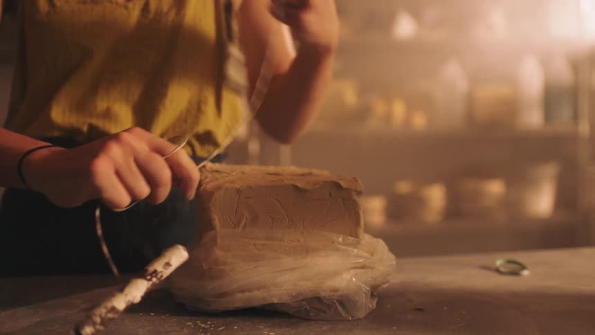 In a warm sunlit studio a woman unwraps a large block of fresh clay and uses a wire cutter to cleanly slice through the dense material to prepare for sculpting