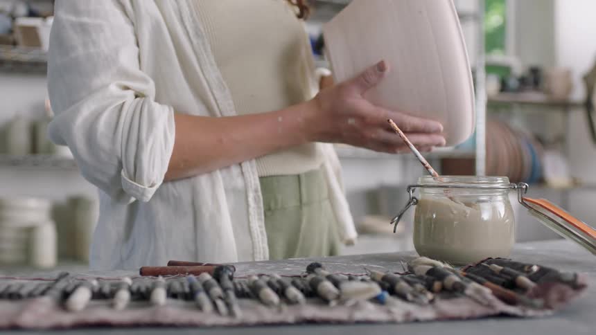 A woman with curly brown hair wearing a white shirt and cardigan opens a jar of tan glaze to paint a large ceramic pot in a brightly lit studio