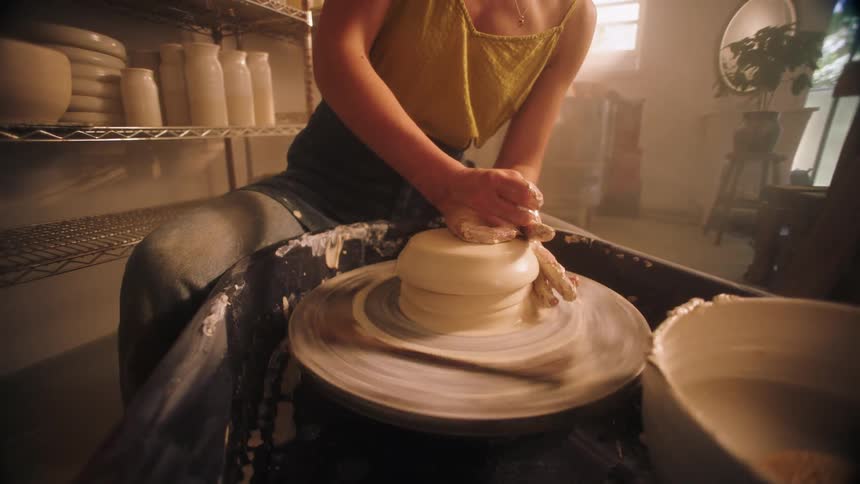 Ceramic artist shapes clay on an electric wheel inside a workshop with shelves of pieces in the background