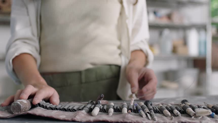 Female potter selects a brush from a rolled bag holding tools in a workshop during the daytime