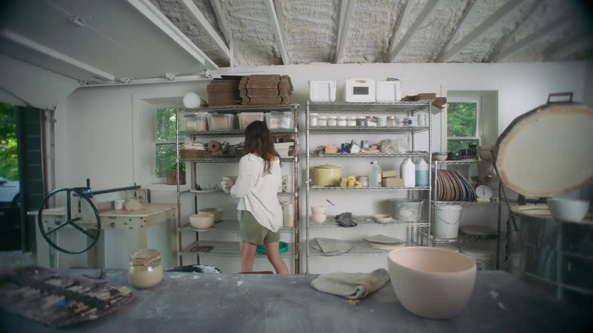 Young adult woman in a pottery studio carrying a ceramic bowl and placing it on a shelf together with many similar items indoors during day time