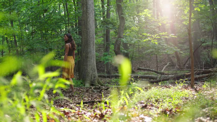 A woman in a yellow skirt slowly walks barefoot through a sun dappled forest looking over her shoulder