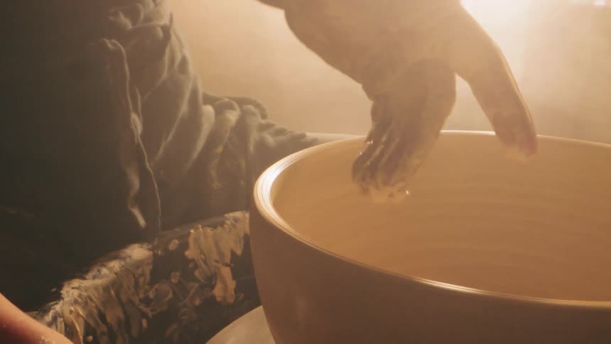 Claycovered hands gently press a wet sponge against the walls of a rotating bowl the motion illuminated by a bright hazy backlight