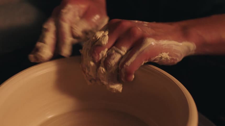A persons arm shaping clay into a pot on a potters wheel inside a studio