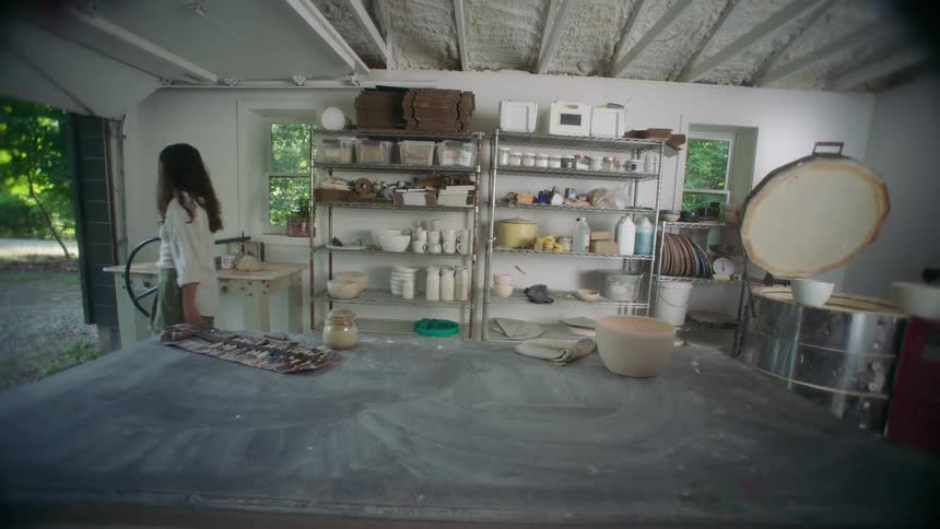 Female potter carries a bowl from the kiln to a shelf before leaving her workshop on a summer day