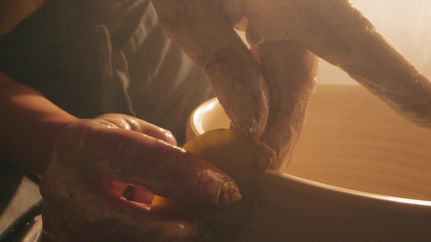 A closeup shot focuses on a persons wet claycovered hands using a small yellow tool to sculpt the interior of a spinning ceramic bowl on a pottery wheel shaping the rim and pulling the walls up