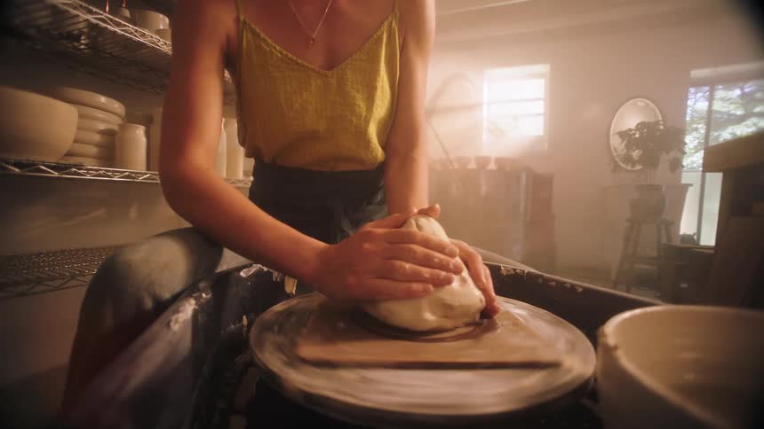In a sundrenched studio a female artisan forcefully throws a lump of raw clay onto the pottery wheel before wetting her hands and skillfully centering the spinning mound