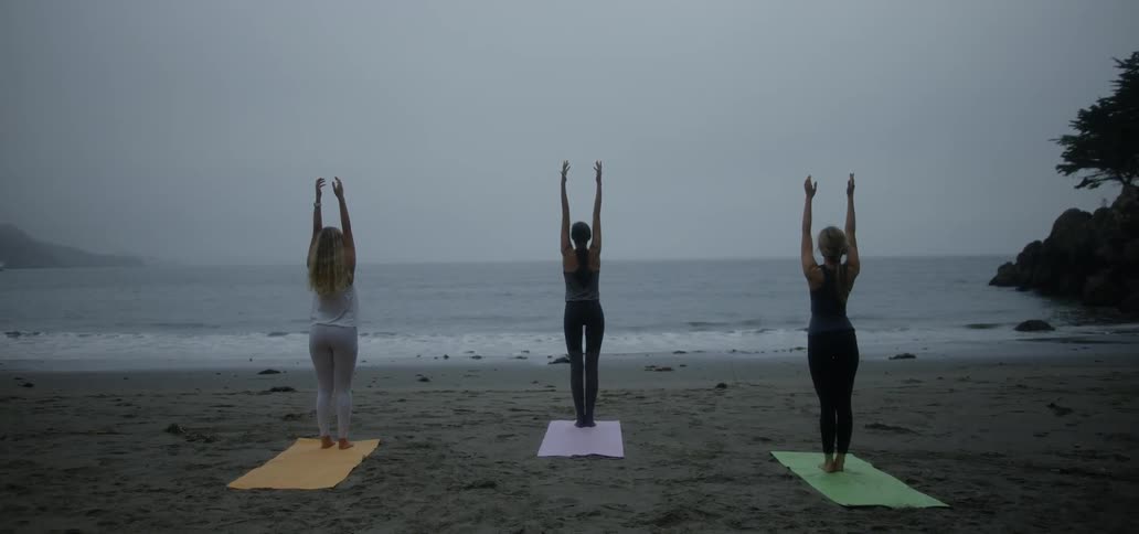 Three women with yoga mats stand on the beach at dusk practicing yoga poses