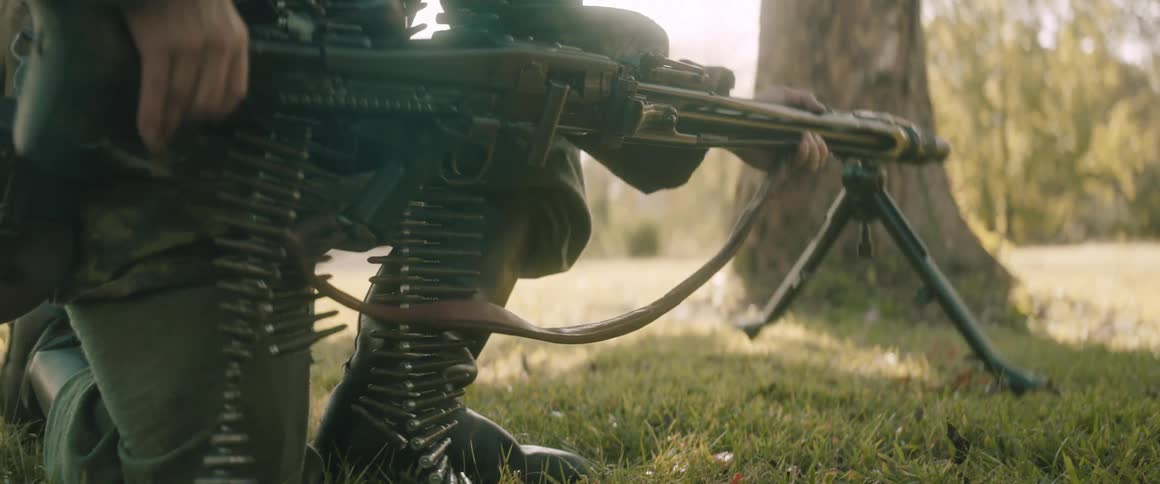 Kneeling soldier operates a machine gun mounted on a bipod on grassy outdoor terrain