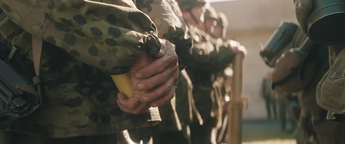 A close view shows the hands of a soldier wearing a speckled camouflage jacket and holding a yellow object standing in a line of soldiers
