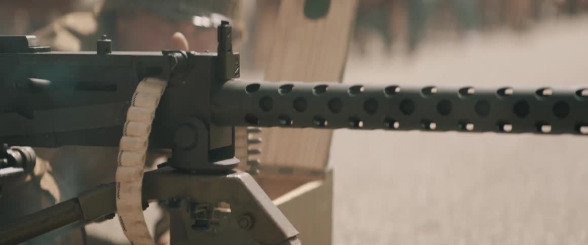 In a detailed closeup a heavy machine gun rattles with rapid fire its perforated cooling jacket vibrating intensely as a cloth ammunition belt feeds steadily into the receiver