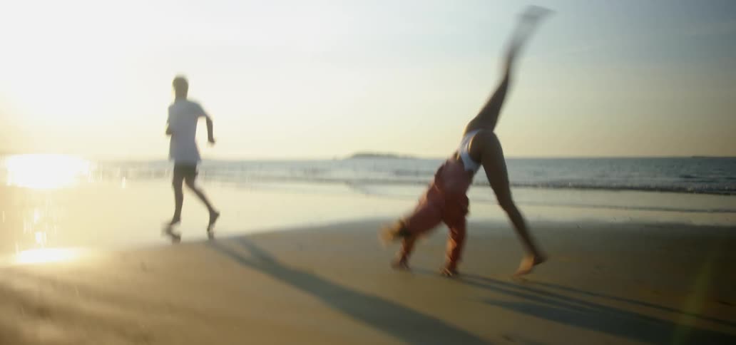 A man and woman run on a beach and then the woman does cartwheels