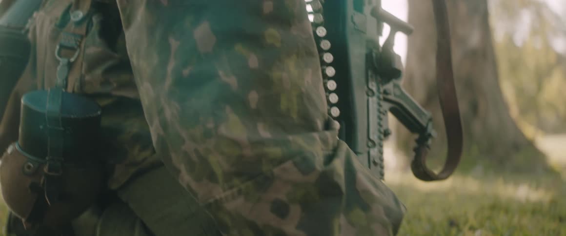A soldier in camouflage draped with belts of ammunition kneels to set up a heavy machine gun on a grassy patch of ground