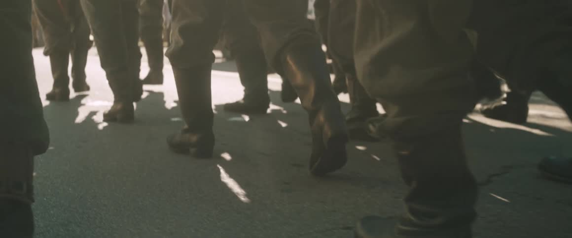 A lowangle closeup captures the rhythmic tread of heavy leather boots striking the pavement highlighting the synchronized movement of the soldiers gaitered legs as they march down a sunlit road