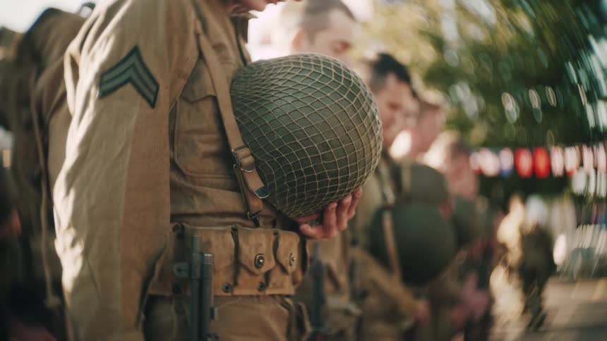 Soldier holds a military helmet while other service members stand in formation in an outdoor setting