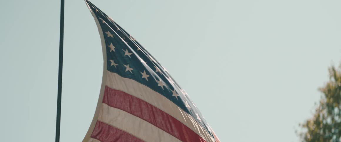 An american flag with slightly muted and aged colors waves on a pole against a bright hazy sky