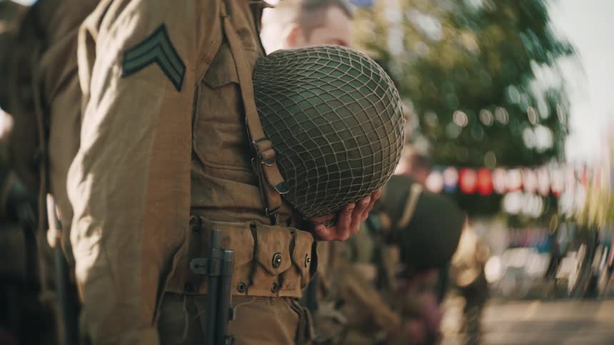 A soldier in a uniform stands outdoors in formation holding his helmet with netting in his left hand with a blurred background of other soldiers and people in an outdoor setting
