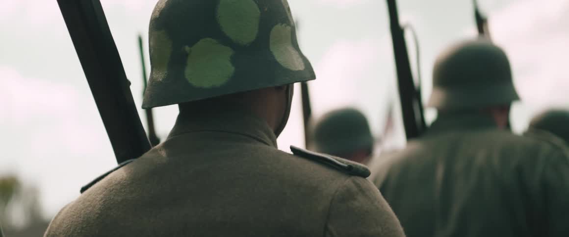 A soldiers back in a gray uniform and a spotted green camouflage helmet is visible while holding a rifle and marching in a line with other soldiers